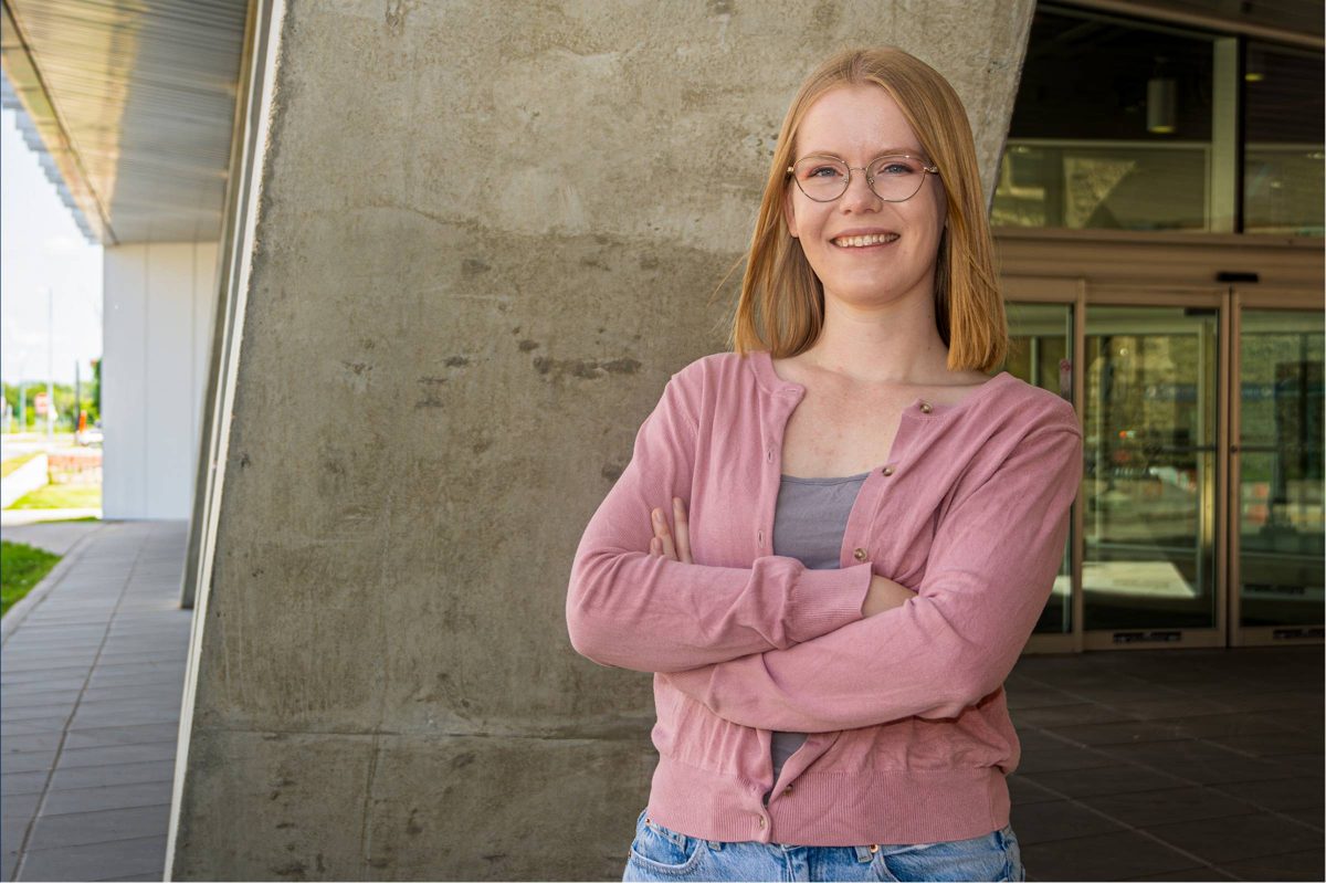 Female wearing glasses standing with arms crossed against a backdrop of a cement wall.