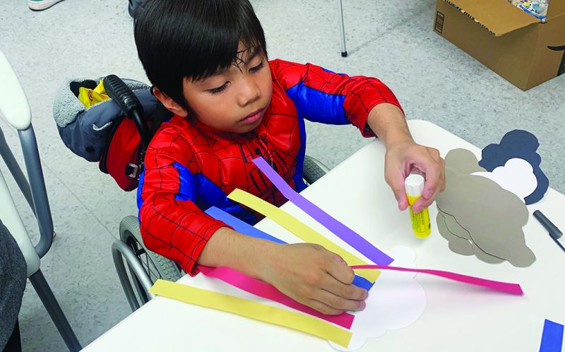 A young boy in a wheelchair, wearing a Spider-man costume, work at a craft table.