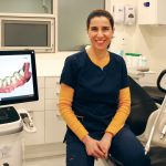 Dr. Hoda Hosseini sits on a dental chair. A 3D image of teeth and gums is on a screen next to her.
