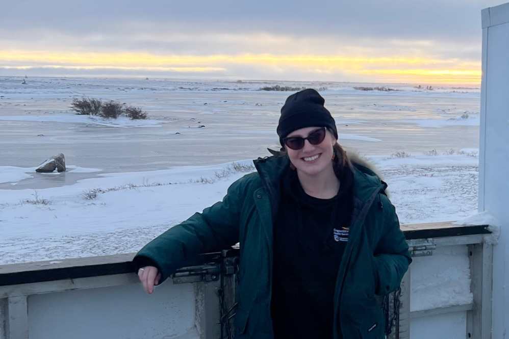 Dr. Emma Anderson poses for a photo wearing winter clothing. A landscape of snow and ice is in the background, with the sun at the horizon.