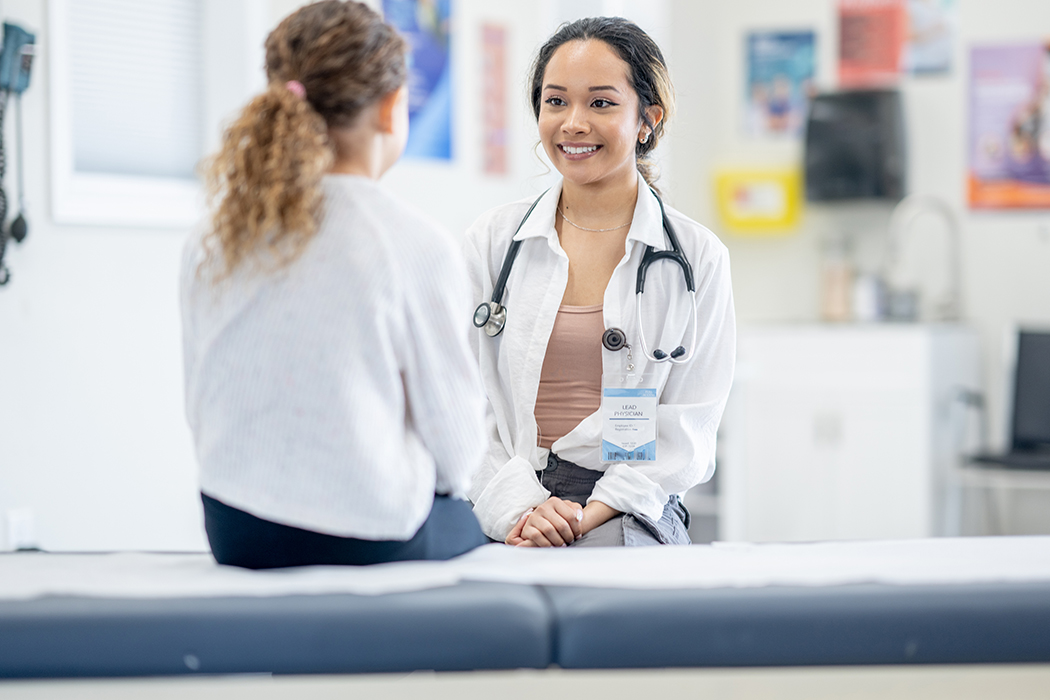 A female doctor talks with a little girl about her health during a medial appointment. She is dressed semi-casually and has a stethoscope around her neck.