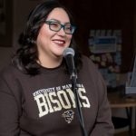 A student with long dark hair and glasses wearing a UM Bisons crewneck sweater stands at a microphone, smiling.