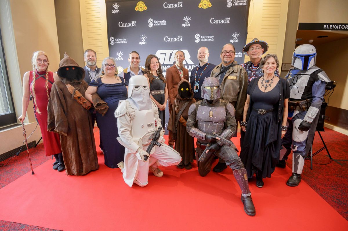 Costumed Star Wars characters pose with voice actors and producers on red carpet in front of Star Wars sign.