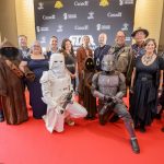 Costumed Star Wars characters pose with voice actors and producers on red carpet in front of Star Wars sign.