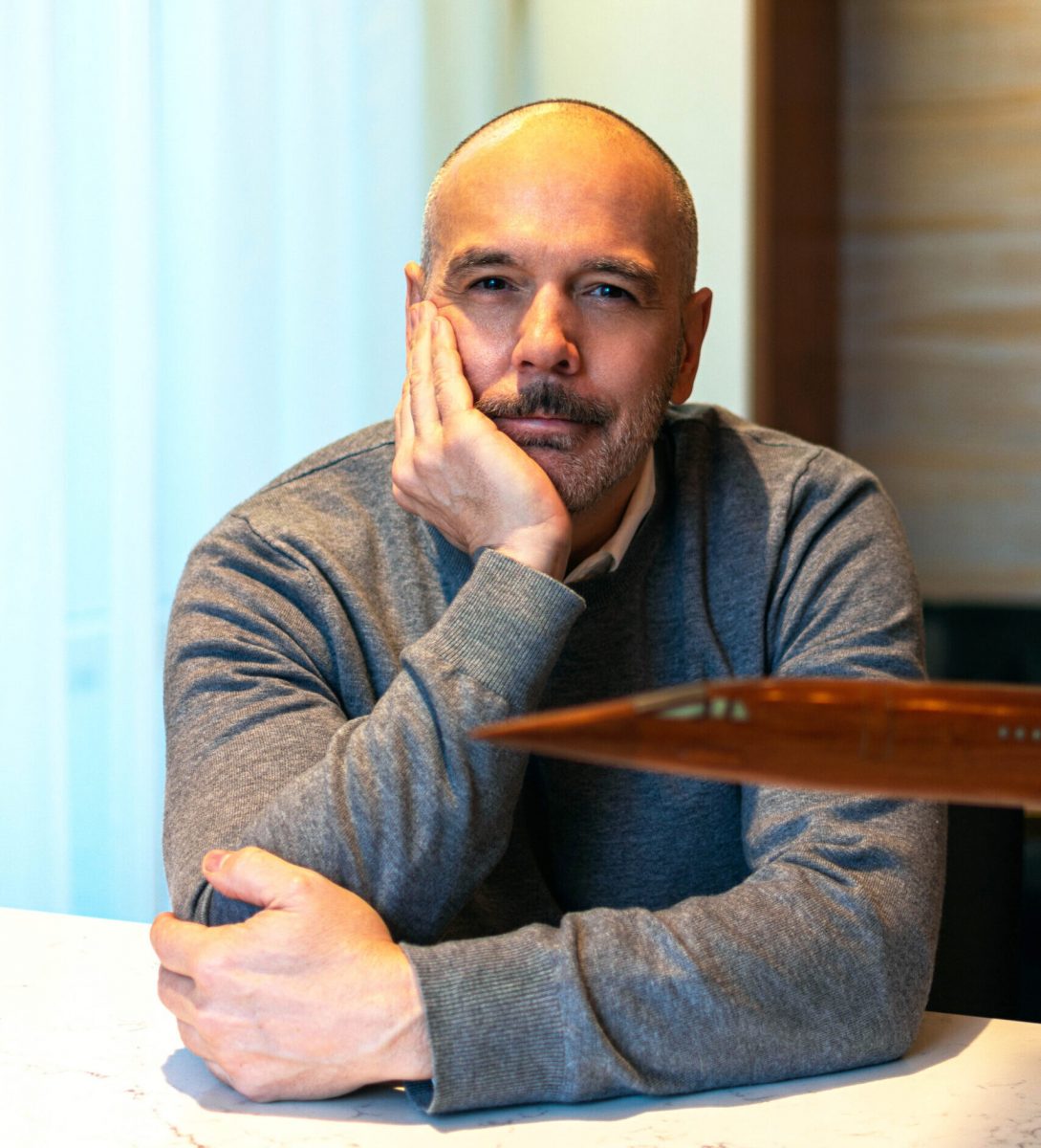 Edmond Huot, UM alum sitting at a desk in a grey sweater with his right hand up to his face, elbows resting on a table