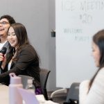 A group of students sit at a table, smiling and listening. One is holding a microphone and there is a whiteboard behind them.