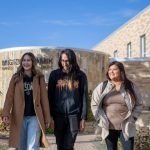 Four Indigenous students walk together outdoors in front of Migizii Agamik - Bald Eagle Lodge at the UM Fort Garry campus.