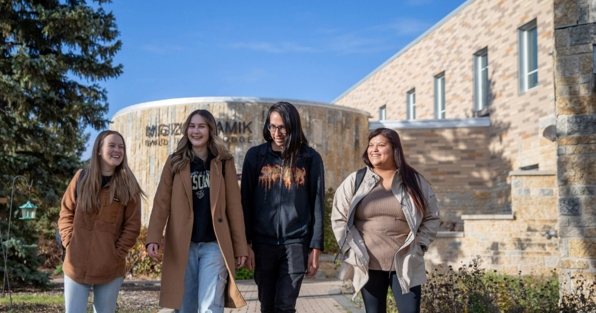 Four Indigenous students walk together outdoors in front of Migizii Agamik - Bald Eagle Lodge at the UM Fort Garry campus.