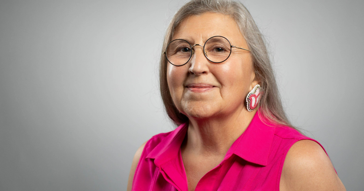 Headshot photo of Pahan PteSanWin. She is wearing glasses, facing the camera and smiling. She has long silver hair and is wearing a pink shirt, set against a grey background.