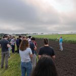 People gather around a speaker on the edge of a tilled field