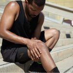 a male athlete sits on steps, holding injured knee