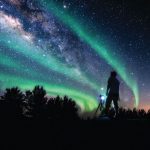 Someone in the foreground has a camera setup on a tripod point up t the sky. Gren and purpl hues of the northerns lights dance infront of an ocean of stars in a dark night sky.