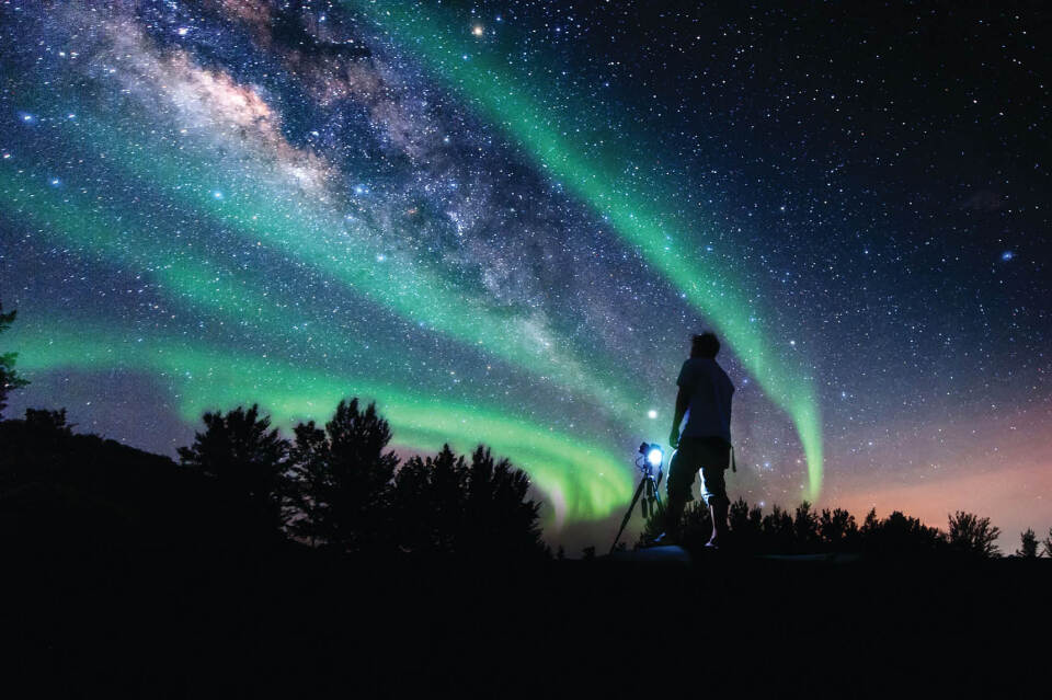 Someone in the foreground has a camera setup on a tripod point up t the sky. Gren and purpl hues of the northerns lights dance infront of an ocean of stars in a dark night sky.