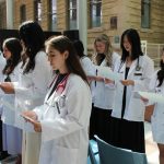 Two rows of students are wearing white coats and stethoscopes. They are standing and each are reading off a paper they are holding.