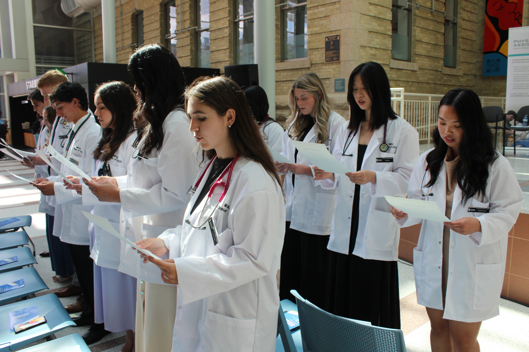 Two rows of students are wearing white coats and stethoscopes. They are standing and each are reading off a paper they are holding.