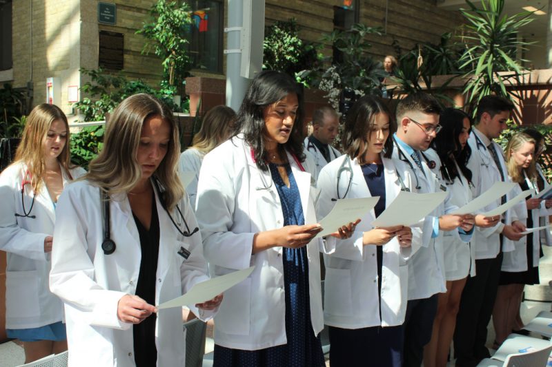 Two rows of students are wearing white coats and stethoscopes. They are standing and each are reading off a paper they are holding.