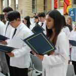 Three rows of students are wearing white coats. They are each holding an open folder and are reading the community code from a paper inside the folder.