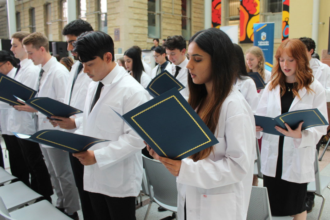 Three rows of students are wearing white coats. They are each holding an open folder and are reading the community code from a paper inside the folder.