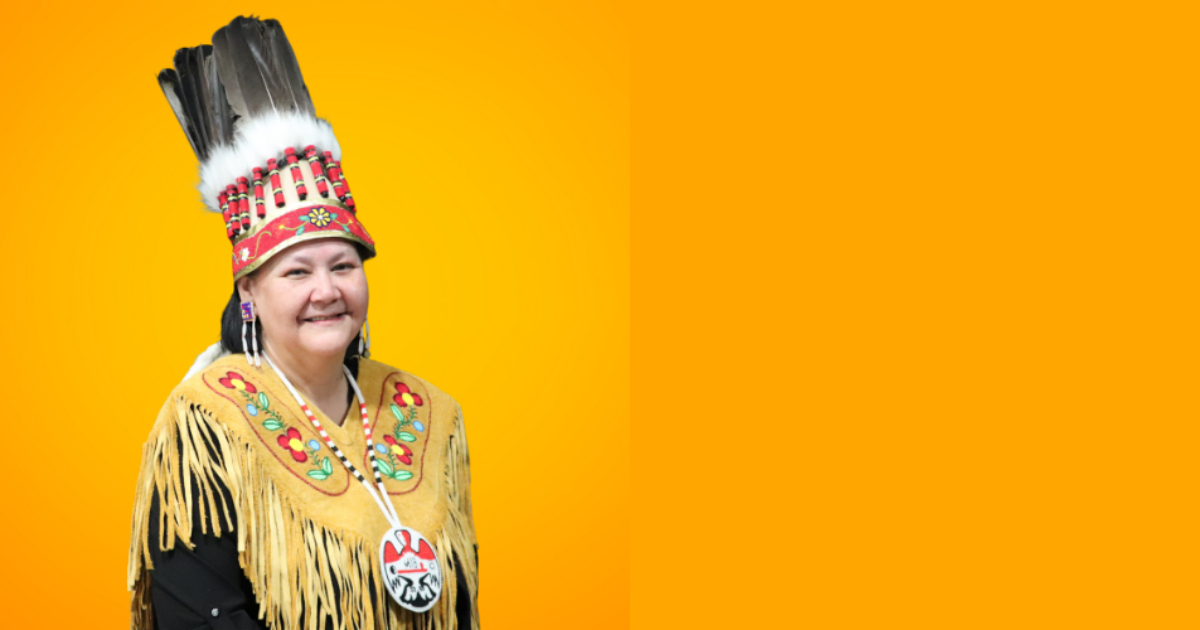 AMC Grand Chief Cathy Merrick dressed in traditional regalia, smiles in front of an orange background.