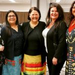 A group of five Indigenous women stand together, smiling at the camera.