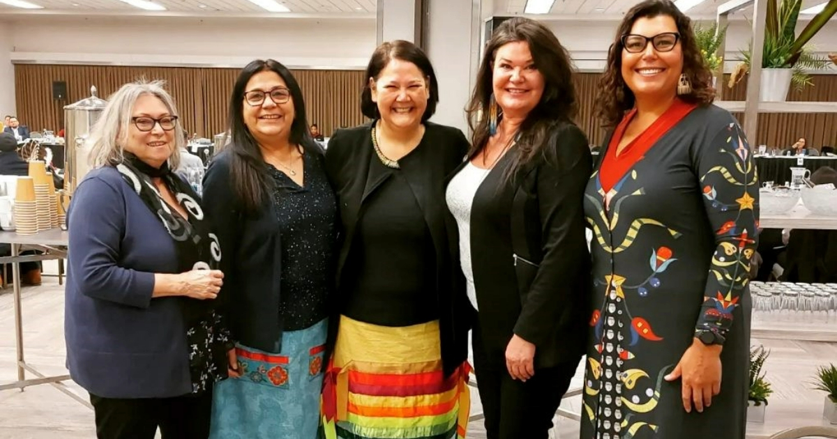 A group of five Indigenous women stand together, smiling at the camera.