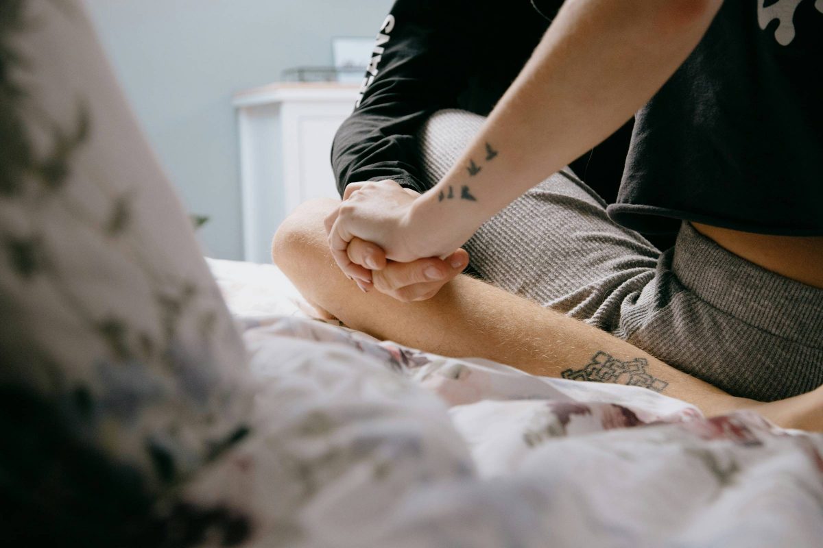 Two people sit cross legged on a bed holding hands. We see only their tattooed arms and legs and the floral bedding.