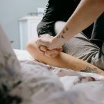 Two people sit cross legged on a bed holding hands. We see only their tattooed arms and legs and the floral bedding.