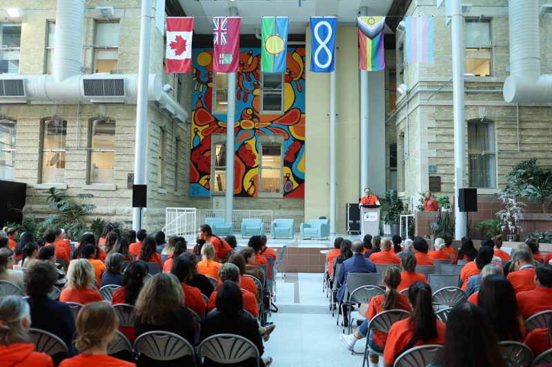 Dr. Nickerson is speaking at a lectern on a stage. He is wearing an orange T-shirt. Audience members are also wearing orange shirts. 