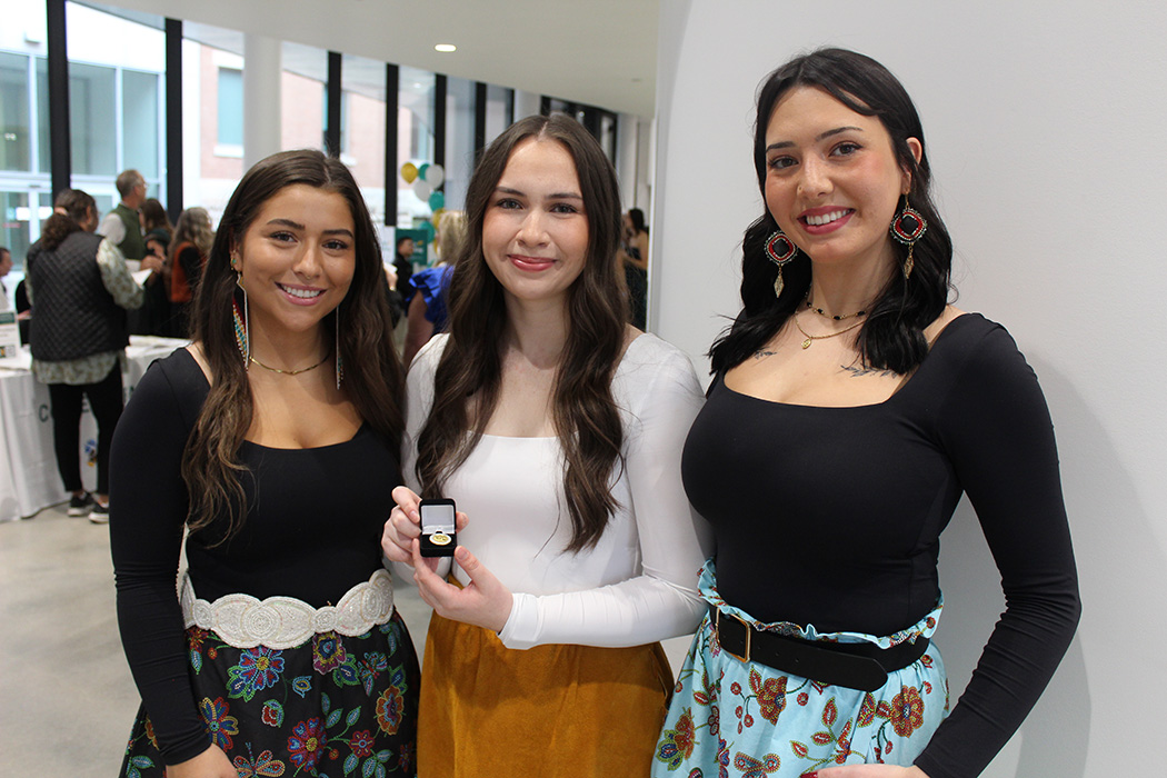 Three female Indigenous students in the lobby of the pinning ceremony. The student in the middle holds her UM nursing pin.