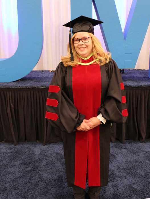 Kristen Valeri wearing a PhD cap and gown in front of a UM sign.