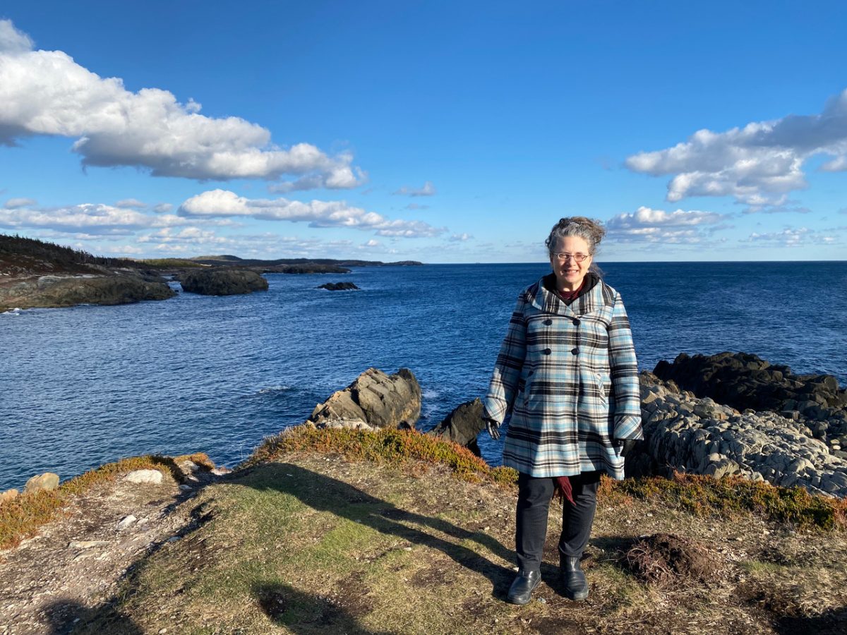 Jody Stark with a plaid jacket standing on a rock that overlooks the ocean - she is smiling at the camera