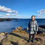 Jody Stark with a plaid jacket standing on a rock that overlooks the ocean - she is smiling at the camera