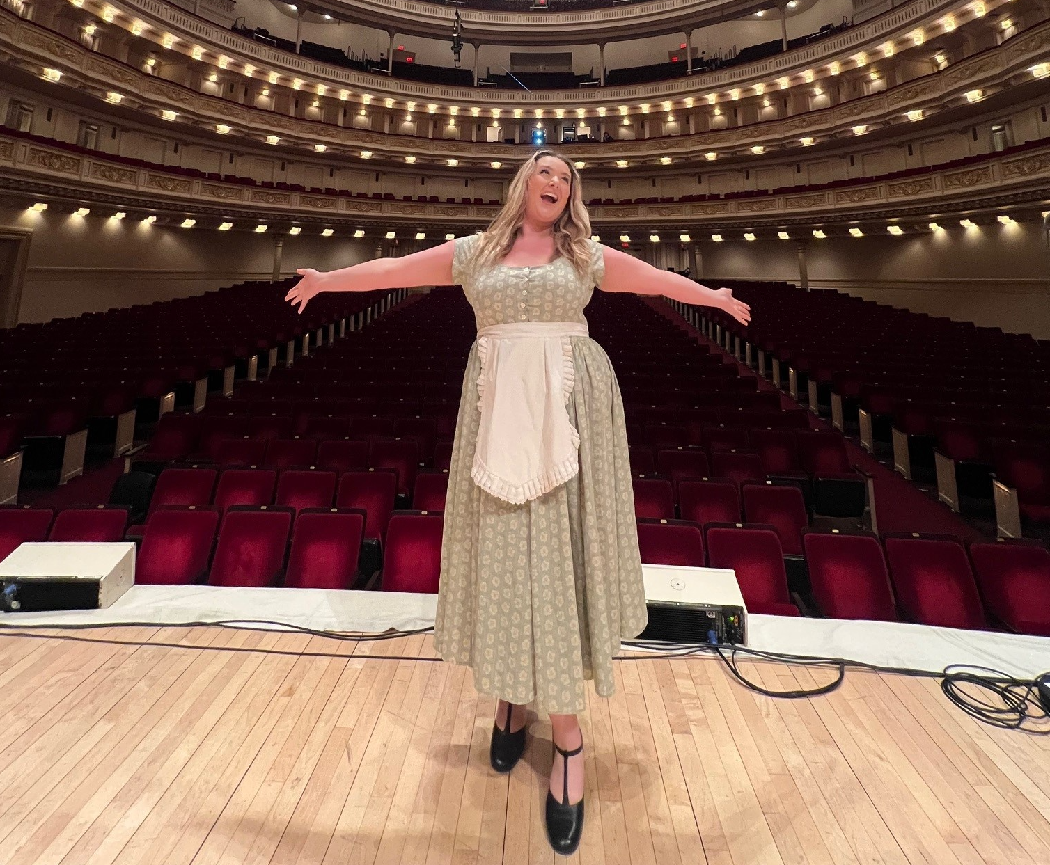 Camryn standing in the theatre on the stage with her arms outstretched with the seats in the background