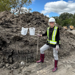 Professor Grace Nickel stands in front of a large mound of locally sourced clay at the Southwood Circle construction site, wearing a hard hat, safety vest, and boots, while holding buckets and a shovel.