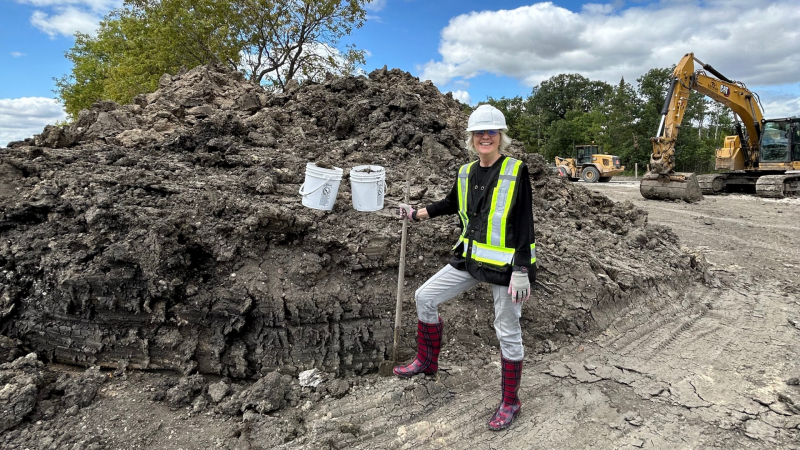 Professor Grace Nickel stands in front of a large mound of locally sourced clay at the Southwood Circle construction site, wearing a hard hat, safety vest, and boots, while holding buckets and a shovel.