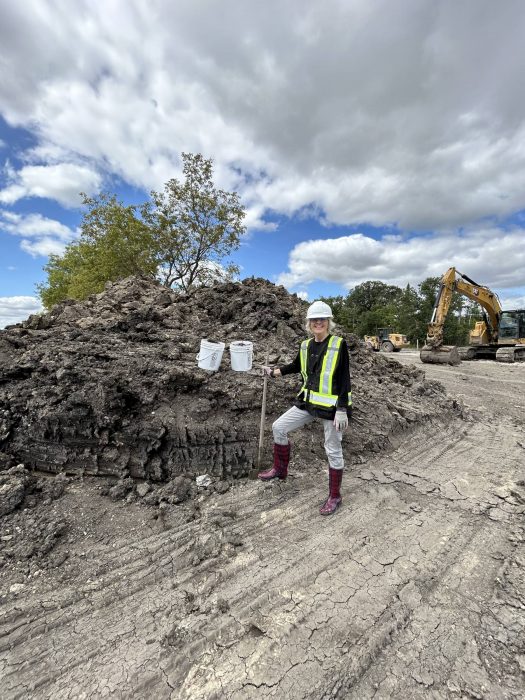 Professor Grace Nickel stands in front of a large mound of locally sourced clay at the Southwood Circle construction site, wearing a hard hat, safety vest, and boots, while holding buckets and a shovel.