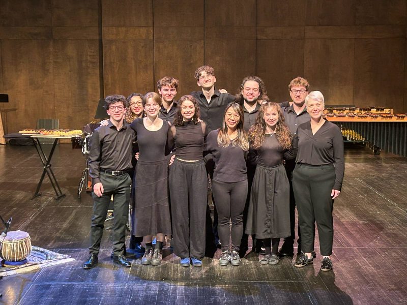 the percussion emsemble stands onstage - they are all dressed in black and smiling and looking at the camera. The names of everyone are listed in the caption from left to right