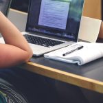 person resting arm on desk with open laptop and notebook