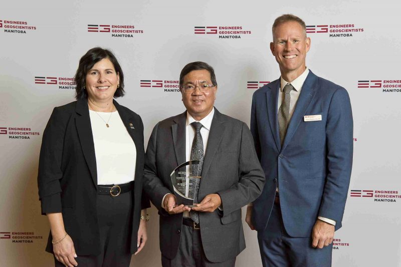 three people posing by EGM backdrop, middle person holding glass award.