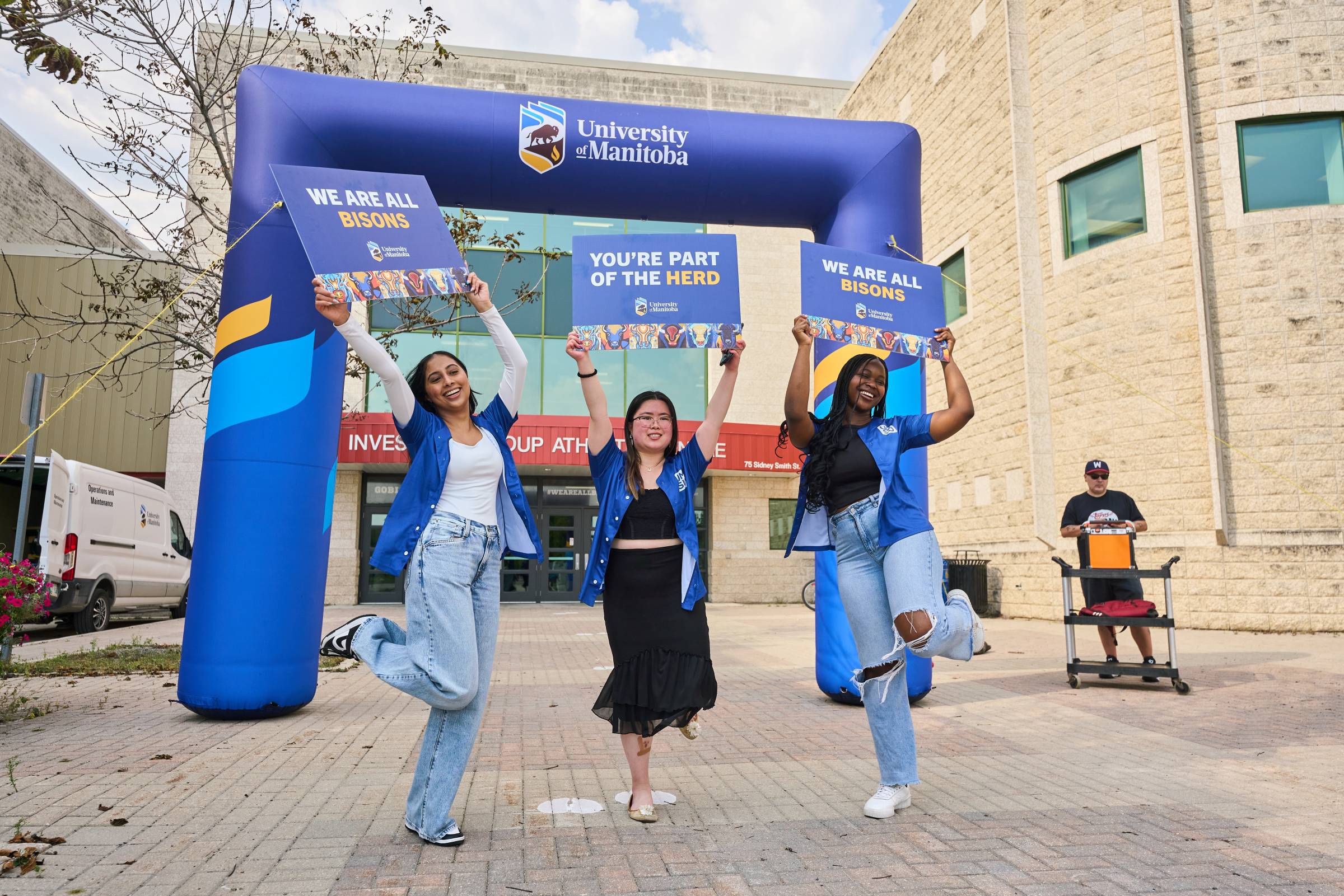 Three student volunteers greet new students at orientation. They stand in front of an inflatable University of Manitoba arch and hold signs that say "We are all Bisons"