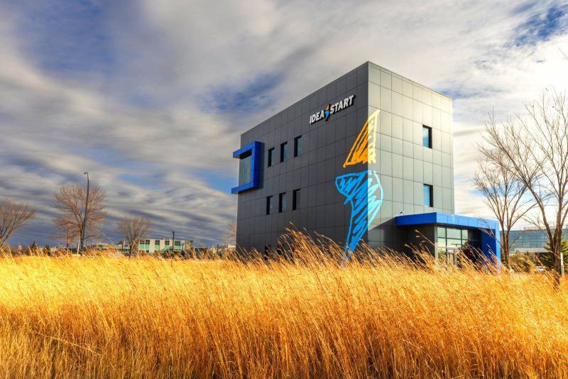 The Idea Start logo features a gold and blue lightening bolt representing the spark of innovation, which now decorates the exterior walls of the Smartpark Event Centre with a field of yellow grass in the foreground.