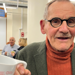 older gentleman in orange turtle neck and grey suit jacket looking at camera holding a coffee mug with red drawing on it.