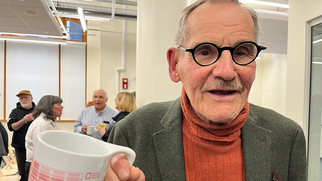 older gentleman in orange turtle neck and grey suit jacket looking at camera holding a coffee mug with red drawing on it.