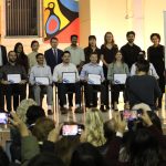 16 people pose for a photo on a stage. A photographer is taking their photo from ground level. Three people in the crowd are taking photos on their phones.