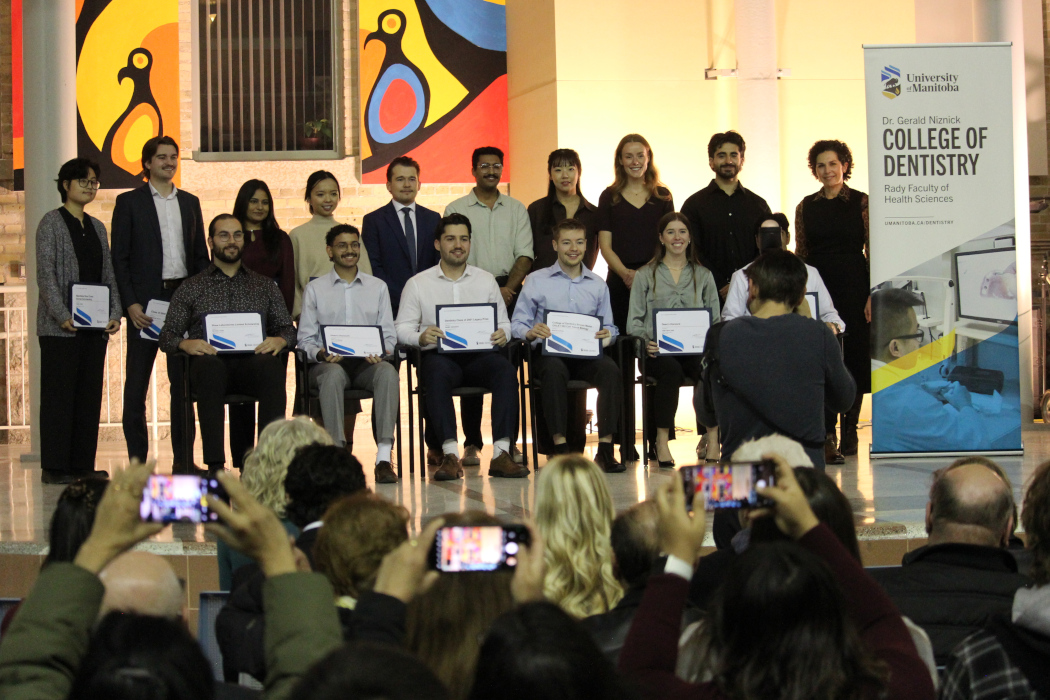 16 people pose for a photo on a stage. A photographer is taking their photo from ground level. Three people in the crowd are taking photos on their phones.