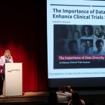 Dr. Laura Saward speaks at a podium beside a projected slide dealing with diversity in clinical trials.