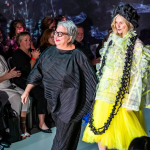 two people walking down a runway. One person wearing all black outfit and one wearing a yellow dress with black very long necklace.