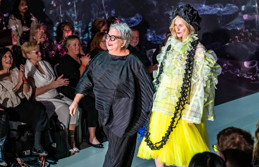 two people walking down a runway. One person wearing all black outfit and one wearing a yellow dress with black very long necklace.