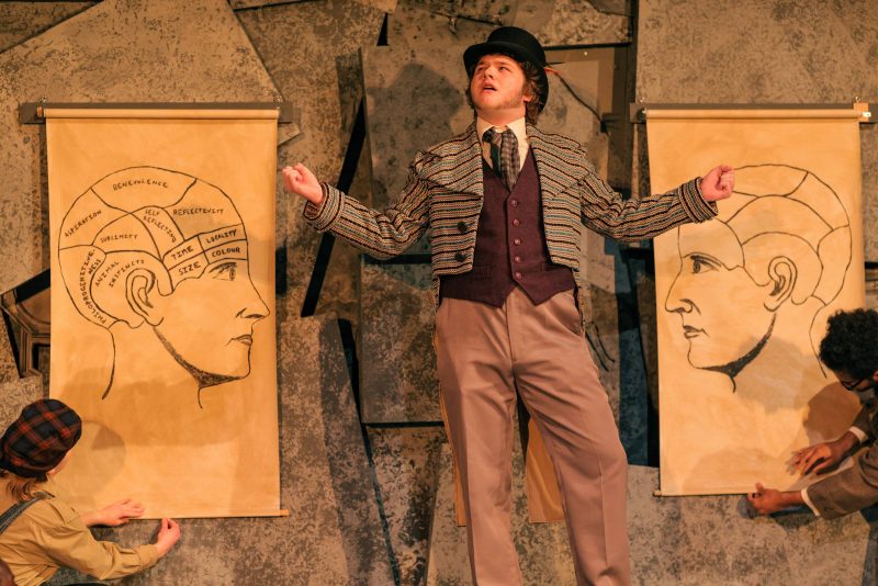 A man dressed from the eighteen hundreds standing between two brain charts hanging on a wall.