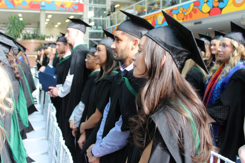 Graduands standing in the atrium at Bannatyne campus for convocation. A colourful piece of Indigenous artwork can be seen in the background encircling them.